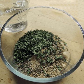 A small glass bowl full of the dried herbs and spices needed to make homamde Italian salad dressing mix on a wooden table with a salad dressing shaker full of vinegar beside it.