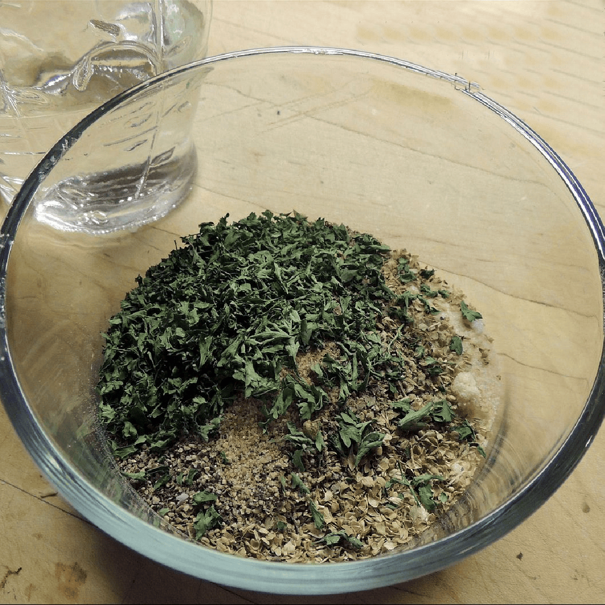 A small glass bowl full of the dried herbs and spices needed to make homamde Italian salad dressing mix on a wooden table with a salad dressing shaker full of vinegar beside it.