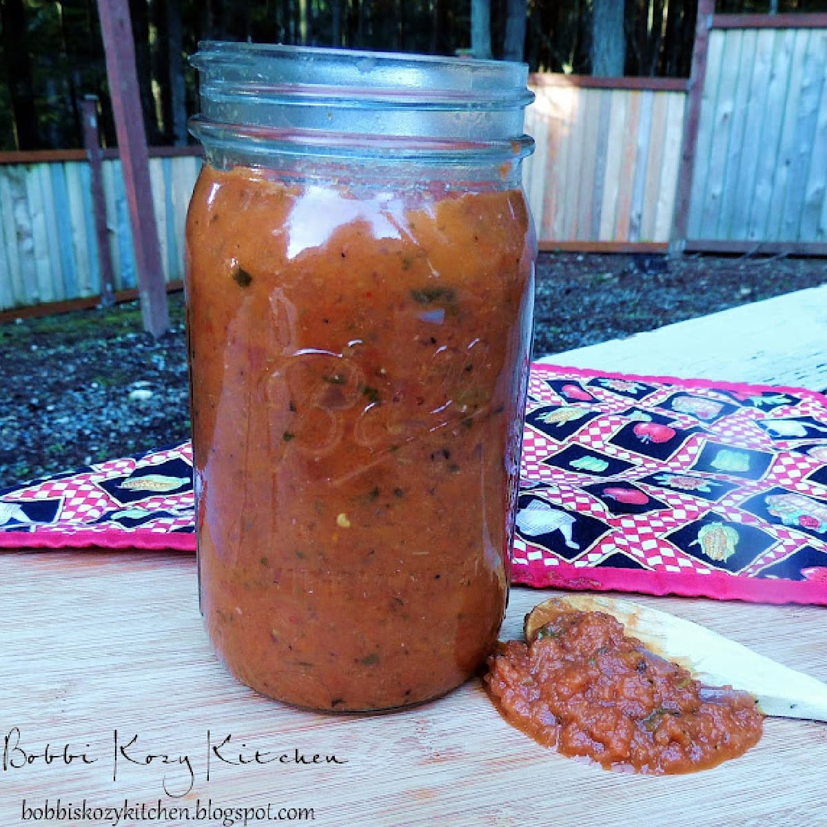 Roasted garlic tomato sauce in a quart mason jar sittin on a wooden cutting board outside with an Italian themed placemat behind it.