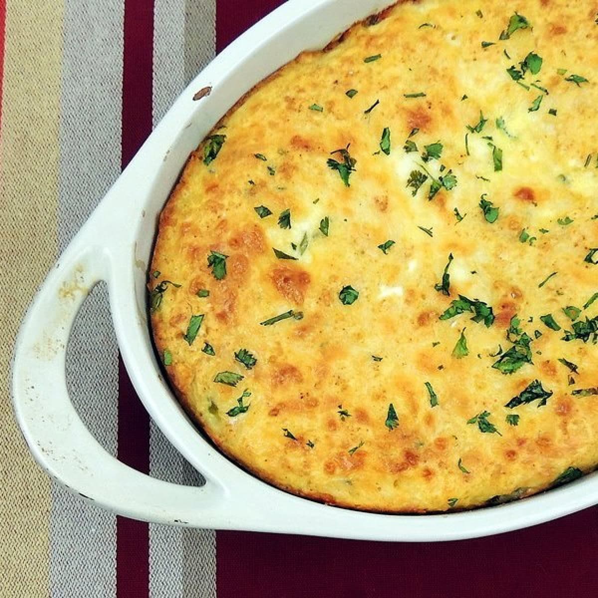 Overhead view of a white baking dish with handles full of Chile Rellenos Casserole with Hatch Green Chiles topped with chopped fresh cilantro. The casserole dish is setting on a earthtone striped placemat.