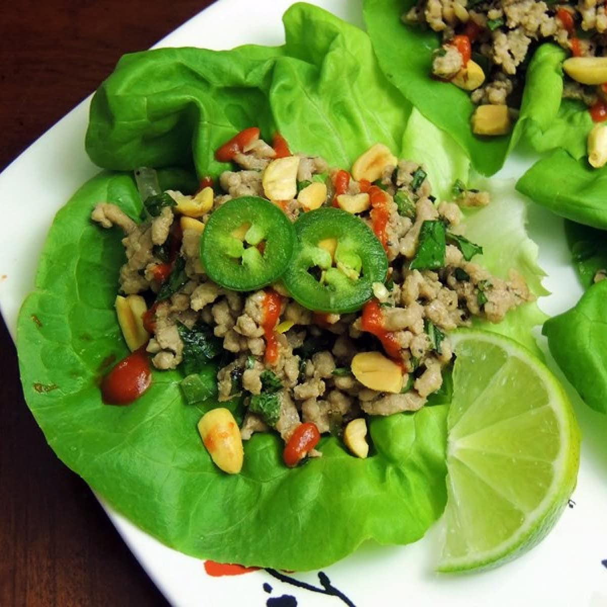 Low-Carb Thai Pork Lettuce Wraps (Larb) on a white plate with Asian decorations. There is a lime wedge on the plate and the plate is on a wooden table.