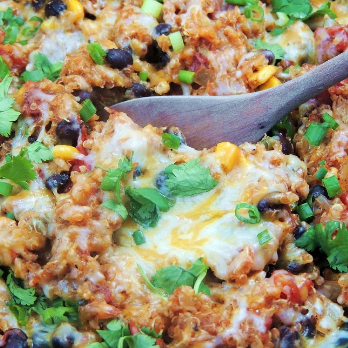 Close-up of Chicken Enchilada Quinoa in a slow cooker, with a wooden spoon scooping it out. The quinoa is topped with melted cheese, fresh chopped green onions, and cilantro.