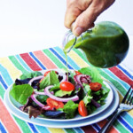 A white male presenting hand pouring homemade basil vinaigrette over a salad on a white plate.