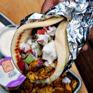 Delicious Sheet Pan Chicken Shawarma wrapped in a pita and topped with raita, held by a hand. The pita is filled with flavorful marinated chicken, accompanied by a refreshing yogurt sauce.