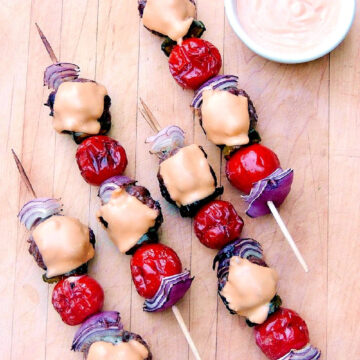 Four cheeseburger kabobs on a wooden cutting board with a small white bowl of deluxe sauce.