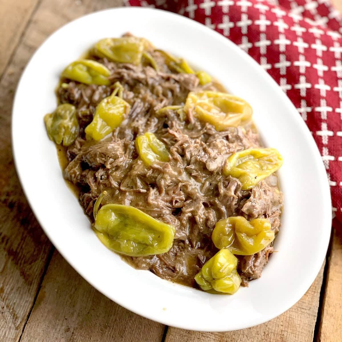 Instant pot Missisippi pot roast shredded in a white serving bowl with pepperoncinis. The bowl is on a wooden table and there is a red and white kitchen towel beside it.