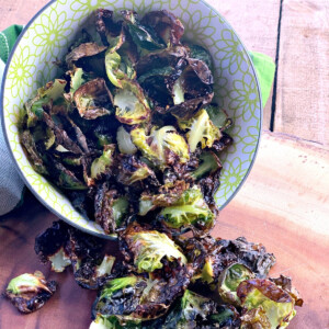 Baked brussels sprout chips in a bowl on it'se side with some chips spilling out onto a wooden cutting board.