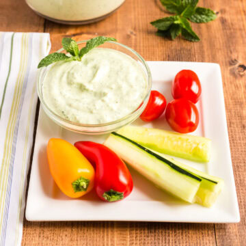 A square white serving tray with mini peppers, cucumber spears, grape tomatoes, and a small glass bowl full of mint raita on a wooden table.