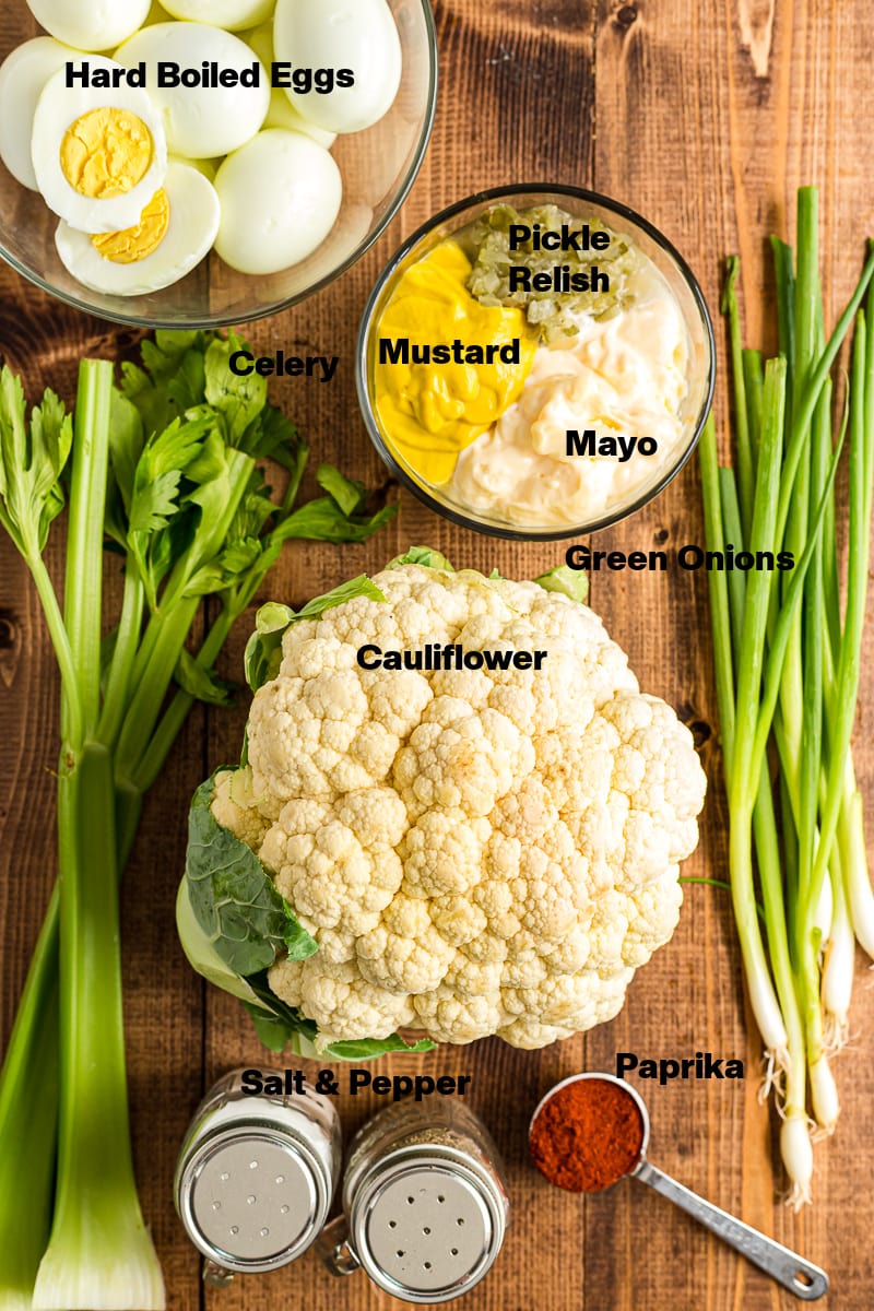 Overhead photo of the labeled ingredients for Deviled Egg Cauliflower Salad, including hard-boiled eggs, cauliflower, green onions, mustard, dill pickle relish, keto mayonnaise, celery, pink salt, cracked black pepper, and paprika, arranged on a wooden table.