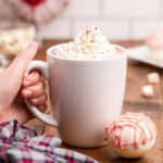 Keto Valentine's Day hot chocolate bomb on a wooden table next to a white mug full of hot cocoa being held by a white female presenting hand.