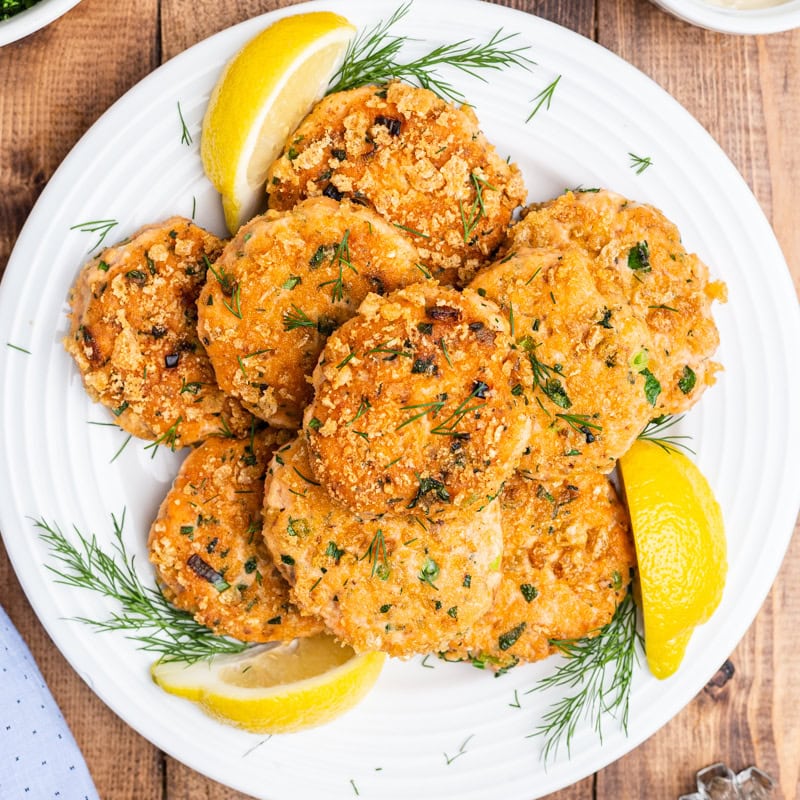 Overhead view of several crispy salmon cakes on a white plate with lemon wedges.