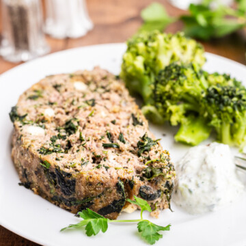 A slice of keto Greek meatloaf on a white plate with tzatziki sauce and broccoli.
