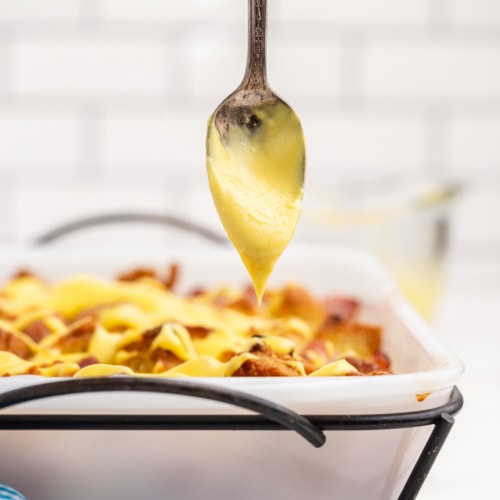 Low carb eggs benedict casserole in a white casserole dish with a silver spoon drizzling hollandaise sauce over the top.