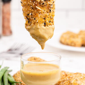 A keto everything bagel chicken tender being dipped into a sugar-free honey mustard. There is a white plate full of the tenders in the background.