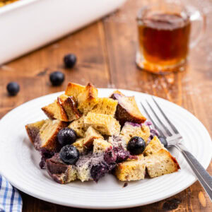 A serving of keto blueberry french toast bake on a white plate with a silver fork on a wooden table. There is a small syrup dispenser behind the plate and blurred in the background is the white casserole dish full of the remaining french toast bake.