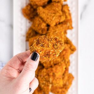 A white female presenting hand holding a keto chicken nugget over a white serving tray full of more keto chicken nuggets below.