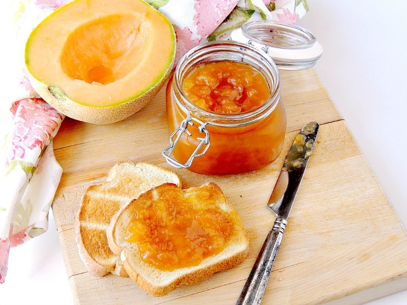 Sugar free Cantaloupe Jelly in a glass jar with a fresh cantaloupe half and toast beside it on a wooden cutting board with a floral towel.
