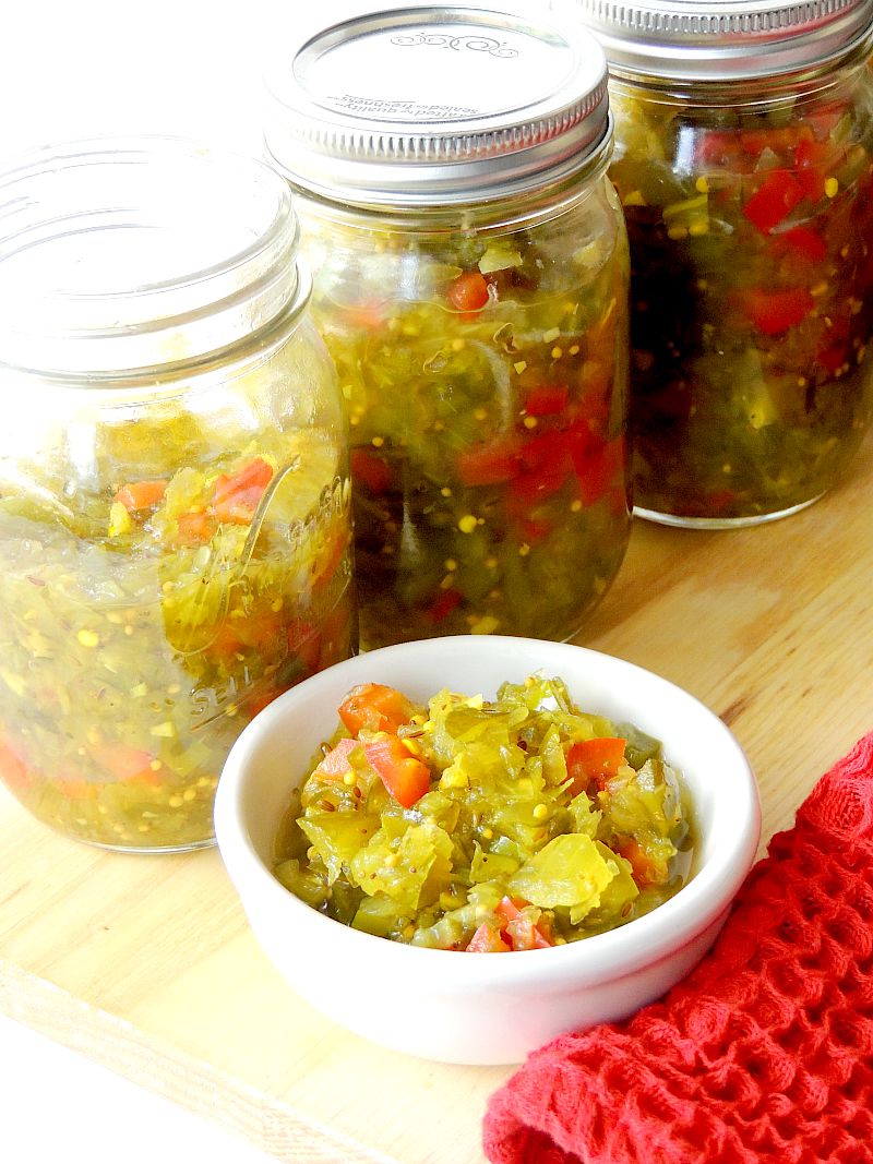 Three pint mason jars full of spicy pickle relish with a small white bowl full next to them on a wooden cutting board.