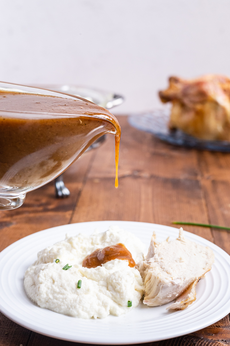 Photo of Perfect Keto Gravy being poured from a glass gravy boat over mashed cauliflower onto a white plate.