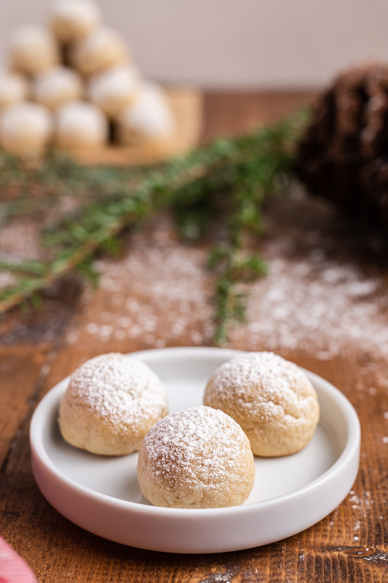 Photo of Keto Snowball Cookies on a small white plate with evergreen branches and more cookies in the background on a wooden table.