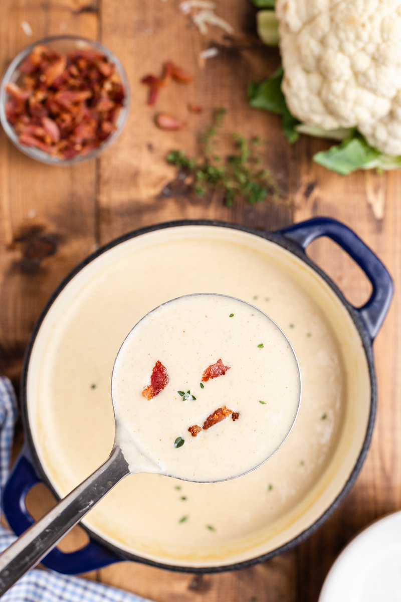 Overhead photo of Easy Cauliflower Cheese Soup in a large blue soup pot on a wooden table with a ladle full of the soup closer to the camera.