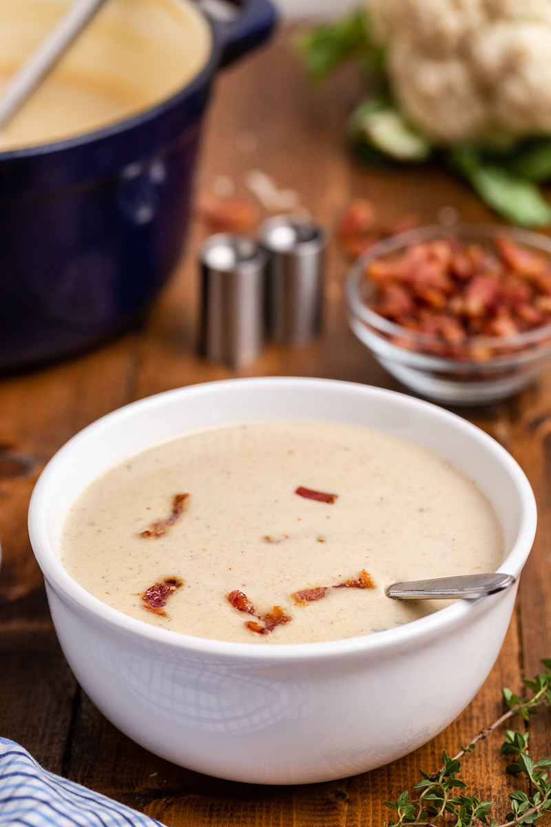 Closeup side view of Easy Cauliflower Cheese Soup in a white bowl with a large blue soup pot in the background on a wooden table.