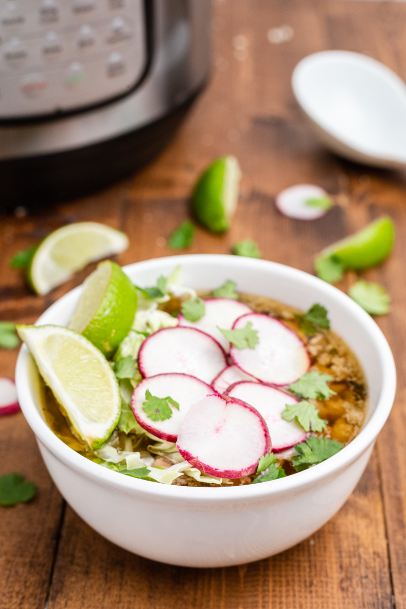 Closeup side view of Keto Pork Pozole Verde in a white bowl on a wooden table with an Instant Pot in the background.