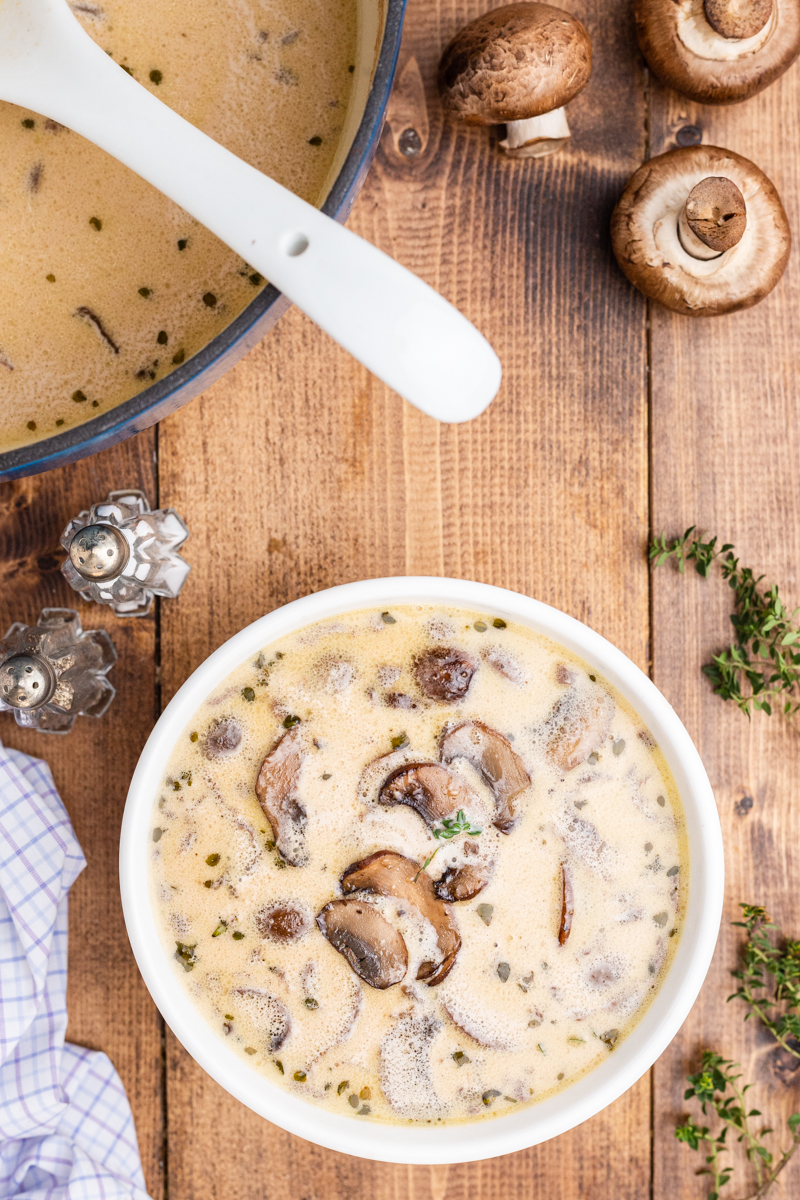 Overhead photo of a white bowl full of Keto Cream of Mushroom Soup on a wooden table with the remaining soup in a large blue soup pot off to the side.