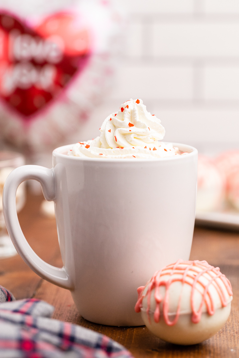 Close up photo of a white mug of hot chocolate with whipped cream and red glitter sprinkle on top and a Keto Valentine Hot Chocolate Bombs sitting next to it on the table.