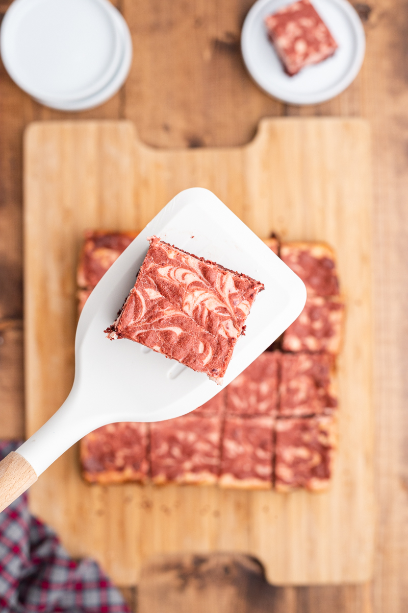 Overhead photo of Keto Red Velvet Cheesecake Brownies cut on a wooden cutting board with one brownie on a spatula closer to the camera.