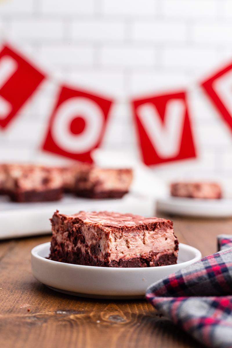 Closeup photo of a Keto Red Velvet Cheesecake Brownie on a white plate with the rest of the batch on a cutting board in the back ground and a sign that says love on the wall.