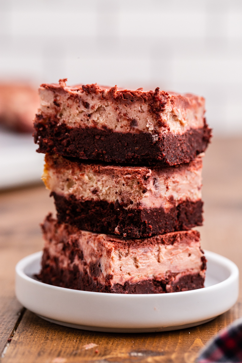 Closeup sideview of 3 Keto Red Velvet Cheesecake Brownies stacked on top of each other on a white plate.