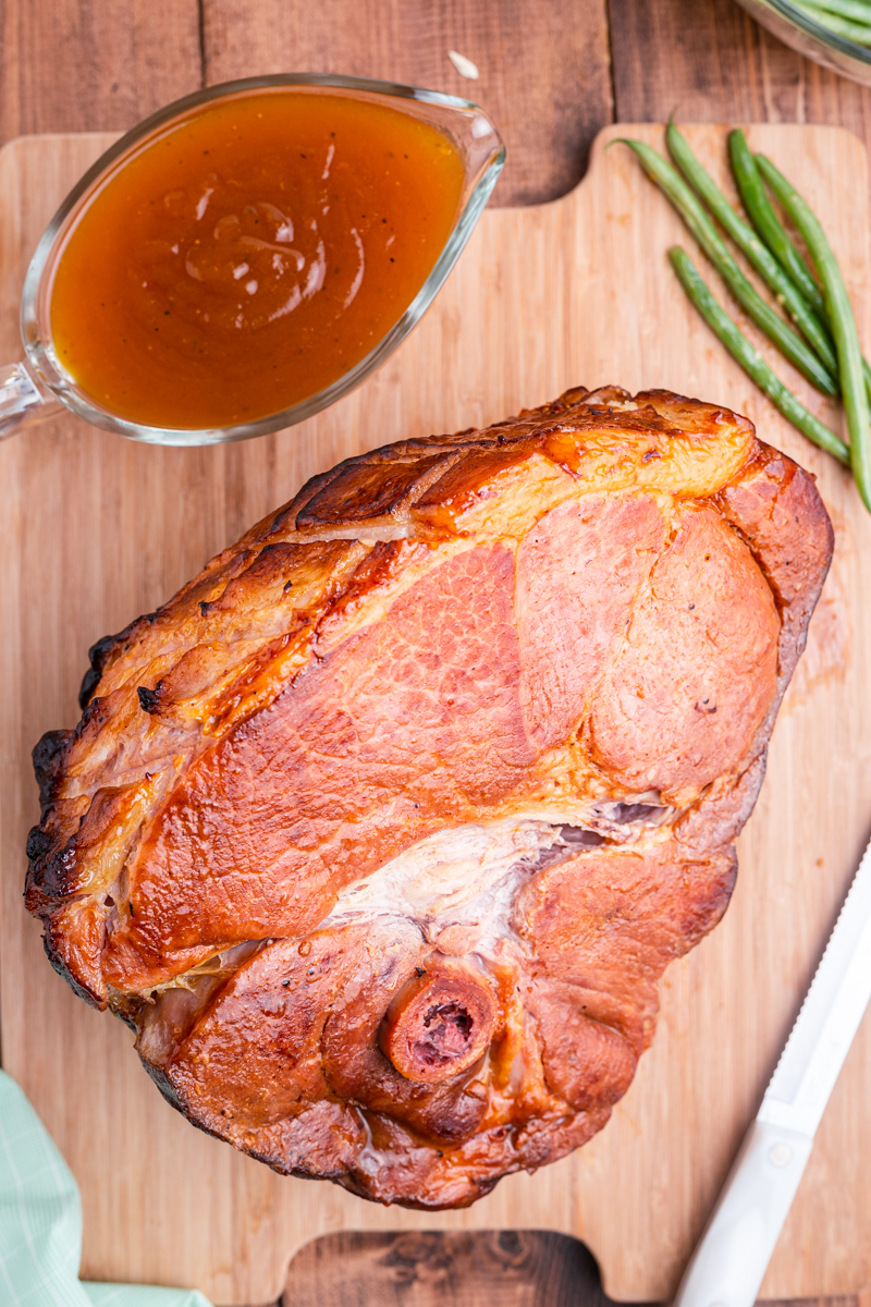 Overhead photo of Keto Apricot Bourbon Glazed Ham on a cutting board with a gravy boat of gravy and green beans next to it.
