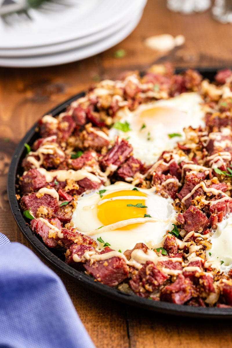 Keto corned beef hash in a cast iron skillet on a wooden table with a stack of white plates behind it.