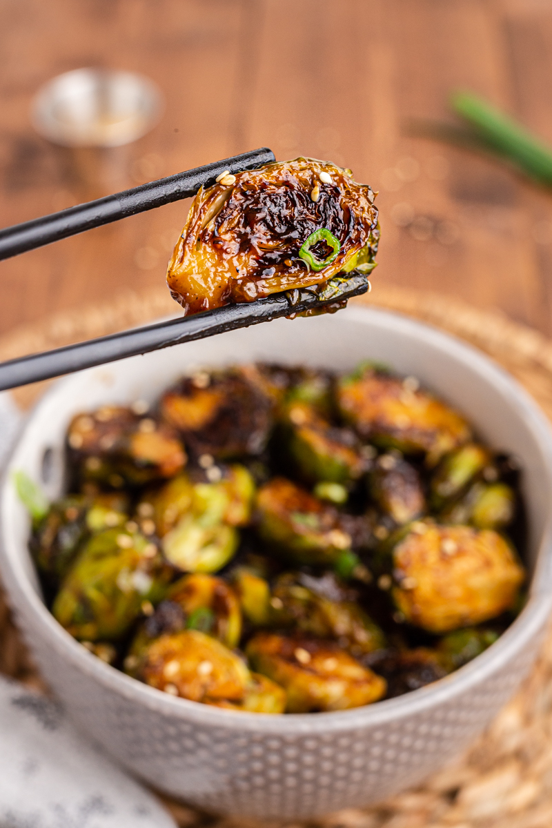 Closeup photo of Kung Pao Brussels Sprouts in a white bowl with someone holding one sprout with chopsticks.