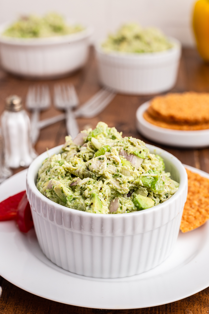 Photo of Keto Avocado Ranch Chicken Salad in a white bowl on a wooden table with crackers and additional bowl of chicken salad in the background.