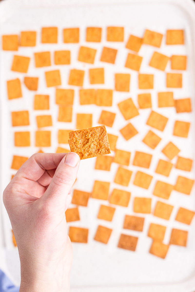 Closeup photo of someone holding a Keto Smoked Cheddar Cracker (Cheez-It) in their hand above a baking sheet covered in the crackers.