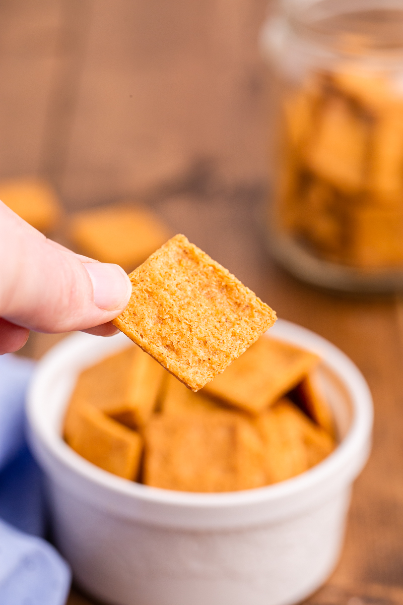 Closeup photo of someone holding a Keto Smoked Cheddar Cracker (Cheez-Its) in their hand with a small white bowl full of them behind their hand.