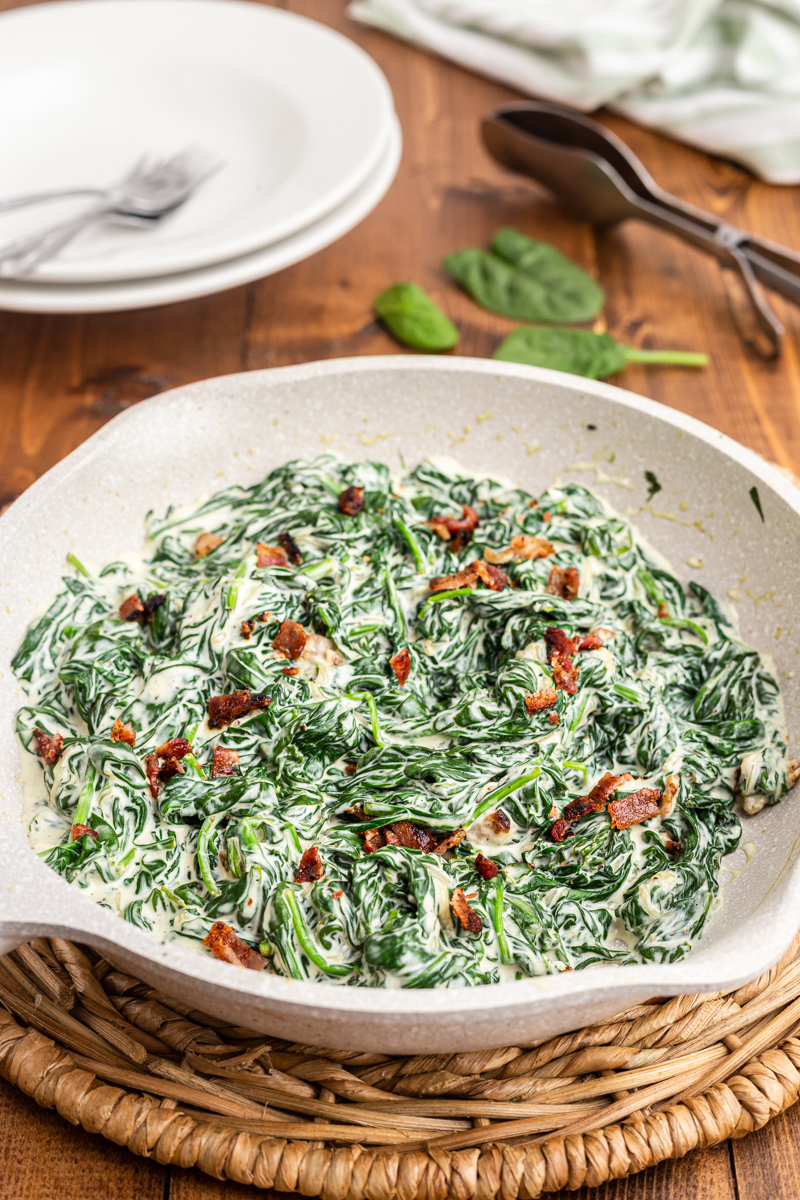 Photo of completed Easy Creamed Spinach recipe in a white skillet on a wooden table.