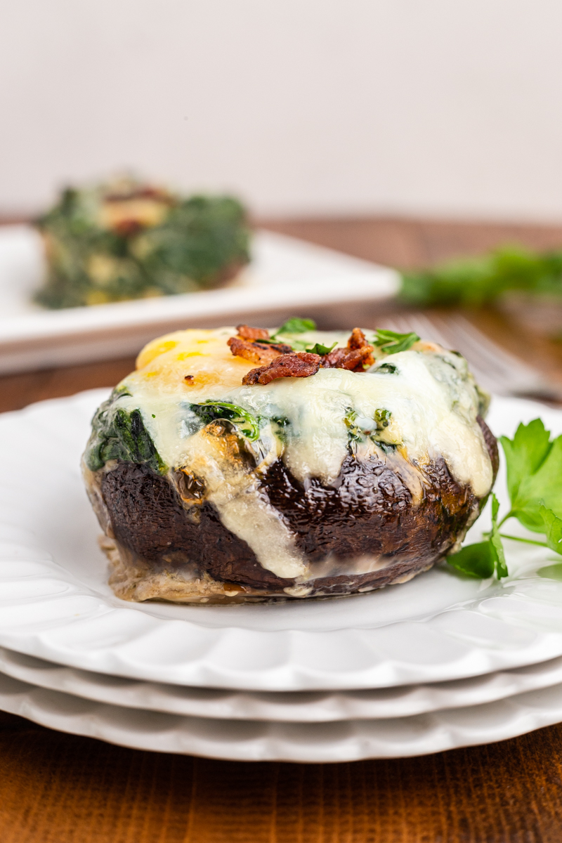 Photo of a Keto Spinach and Egg Stuffed Mushroom on a white plate with a platter of them in the background.