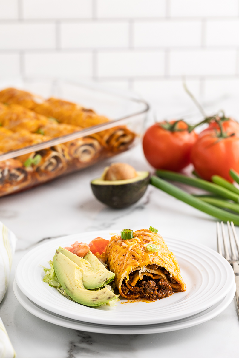 A Low Carb Beef Enchilada Rollup on a white plate with avocado slices and a glass baking dish with more Low Carb Beef Enchilada Rollups in it in the background.