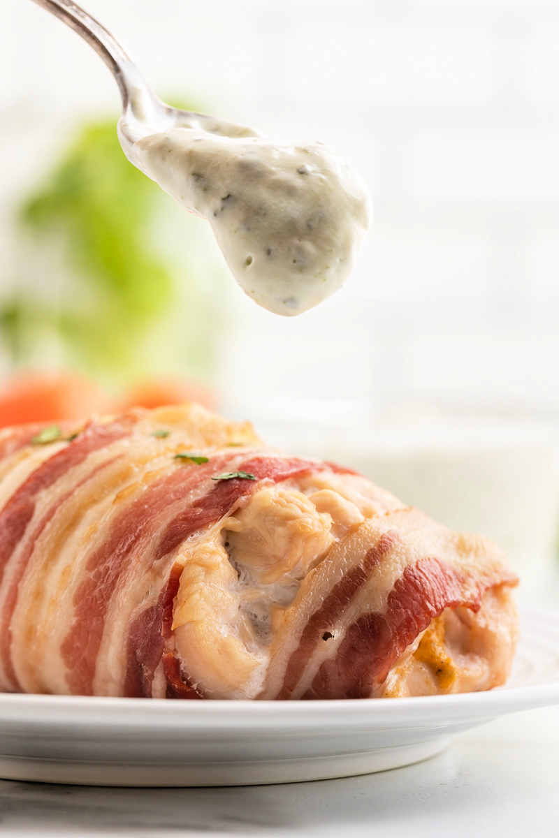 A spoonful of Cilantro Lime Crema being dropped onto a Bacon Wrapped Jalapeno Popper Stuffed Chicken Breast.