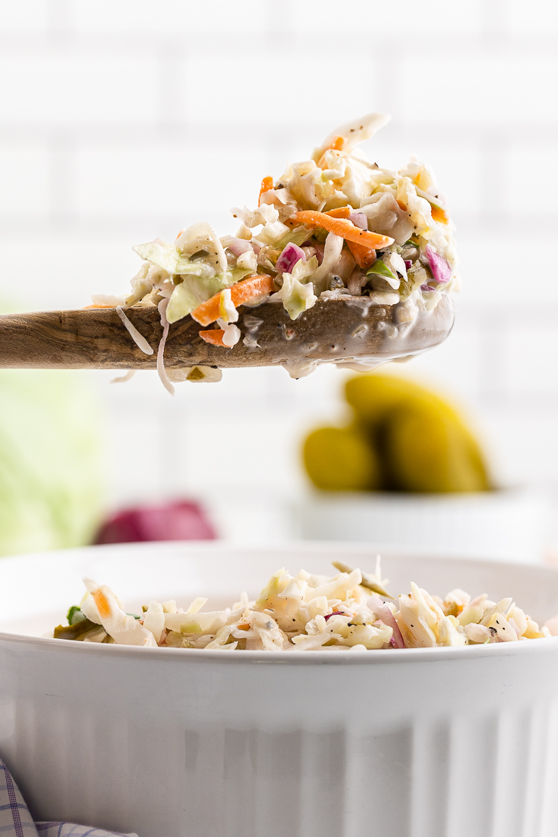 Side view of a white serving dish with Dill Pickle Coleslaw and a serving spoon full above the dish.
