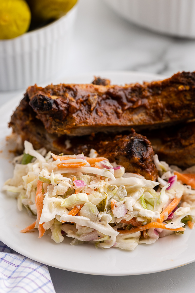 Delicious dill pickle coleslaw served on a white plate alongside mouthwatering BBQ ribs. The plate rests on a white counter, adorned with a blue and purple plaid dish towel.