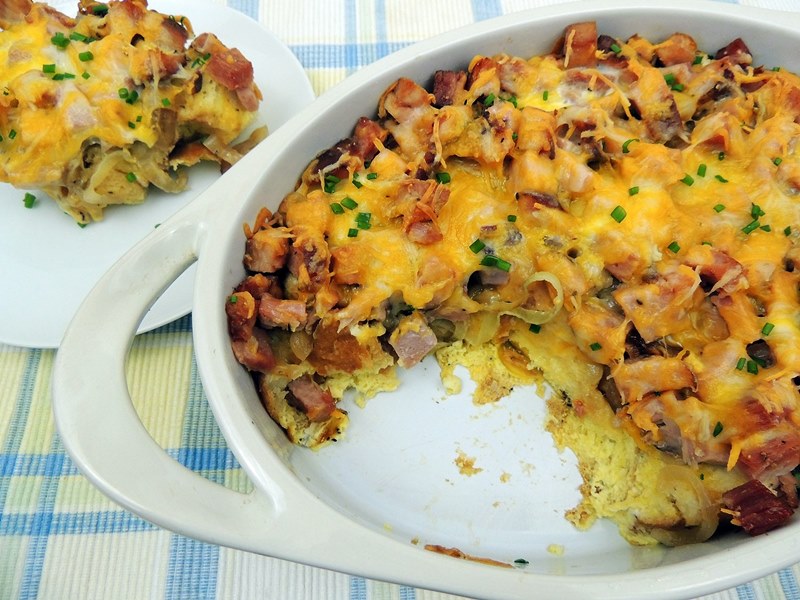 Overhead view of make ahead ham and cheese breakfast casserole in a white casserole dish with one serving on a white plate.