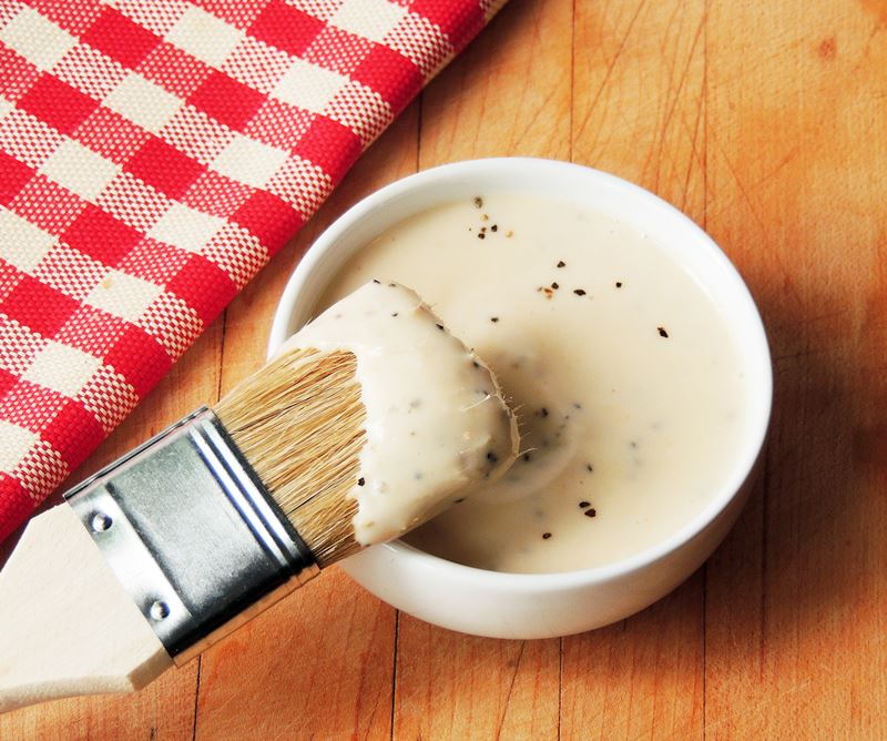A small white bowl full of Alabama white BBQ sauce with a basting brush resting on the edge.