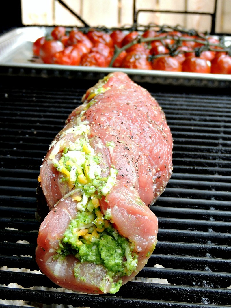 Broccoli Pesto and Cheese Stuffed Grilled Pork Tenderloin cooking on a charcoal grill with cherry tomatoes behind.