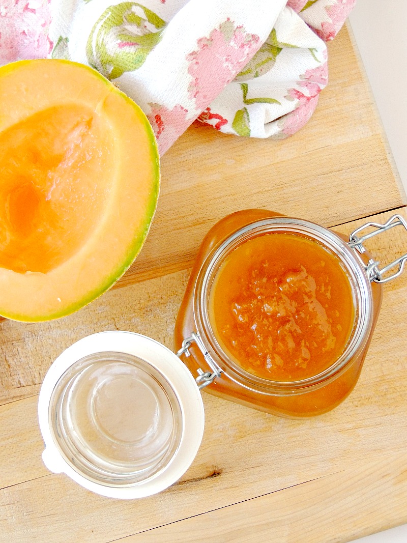 Cantaloupe Jelly in a glass jar with a fresh cantaloupe half beside it on a wooden cutting board with a floral towel.