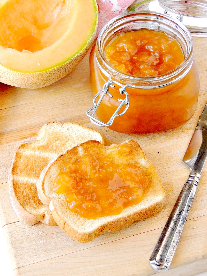 Sugar free Cantaloupe Jelly in a glass jar with a fresh cantaloupe half and toast beside it on a wooden cutting board.
