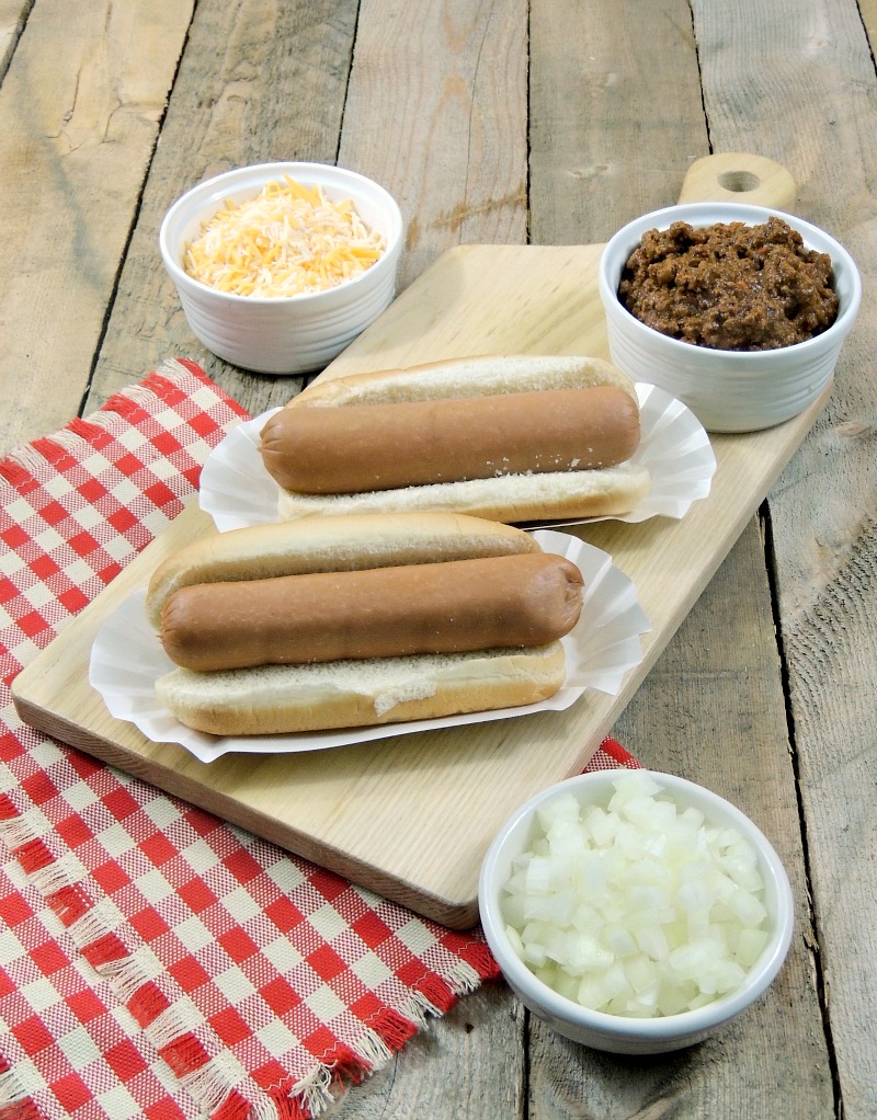 2 plain hot dogs on buns on a cutting board with bowls of chopped onions and shredded cheese on a wooden background with a red and white checkered napkin.